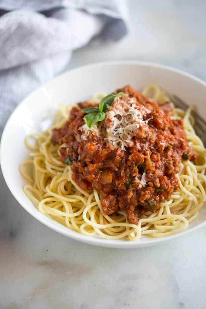 A white bowl full of spaghetti noodles with homemade spaghetti sauce on top and fresh grated parmesan cheese and basil.