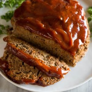 A loaf of meatloaf on a white plate with sauce on top and two slices cut from it, with fresh parsley in the background.