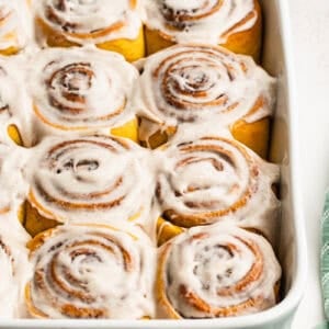 A homemade Pumpkin Cinnamon Rolls recipe in a white baking dish with a cinnamon cream cheese frosting spread on top.