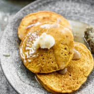 Three homemade Pumpkin Pancakes stacked on top of each other with a spoonful of butter on top and syrup drizzled.