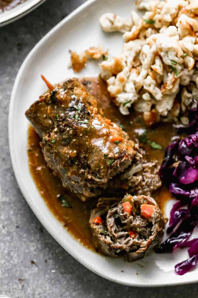 German Rouladen served on a plate with a bite taken out to show the carrot, pickle and onion inside, next to red cabbage and spaetzle.