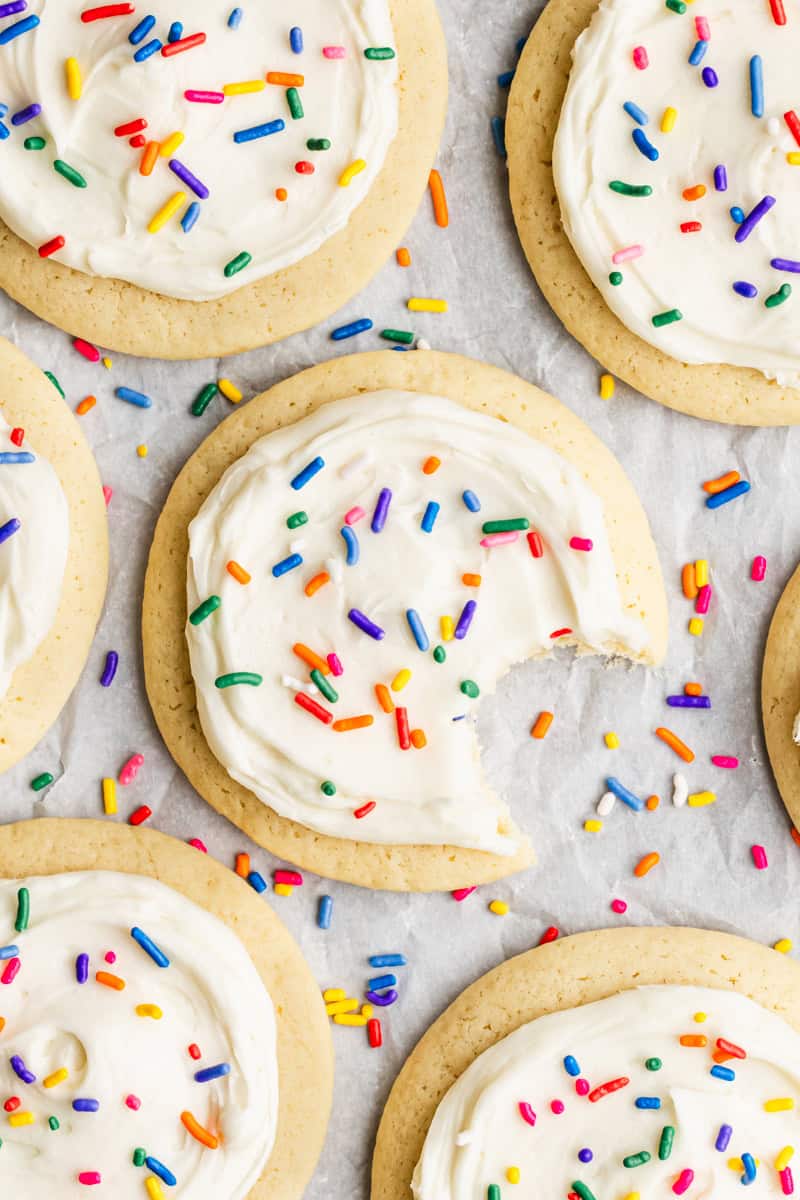 A soft sugar cookie recipe showing the cookies on a piece of parchment paper, frosted with cream cheese frosting and topped with a pinch of rainbow sprinkles. 