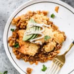 A serving of homemade Tamale Pie on a white plate, topped with fresh avocado slices, sour cream, and salt and pepper.