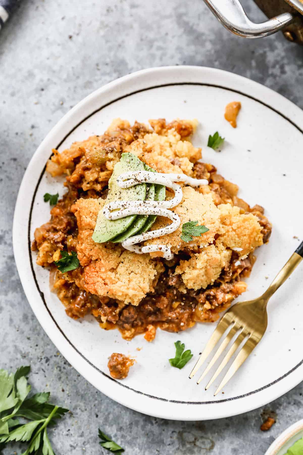 A piece of homemade Tamale Pie on a white plate topped with fresh avocado, sour cream, and salt and pepper. 