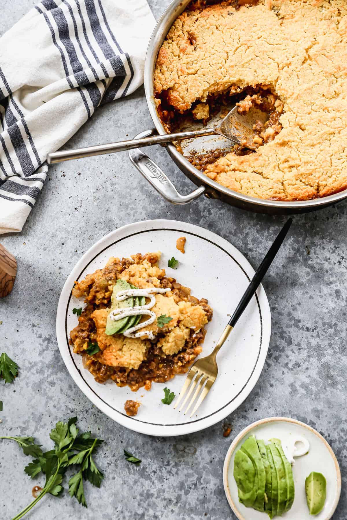 A plate of Mexican Tamale Pie with avocado and sour cream on top, next to the pan of Tamale Pie and a small plate of sliced avocados. 