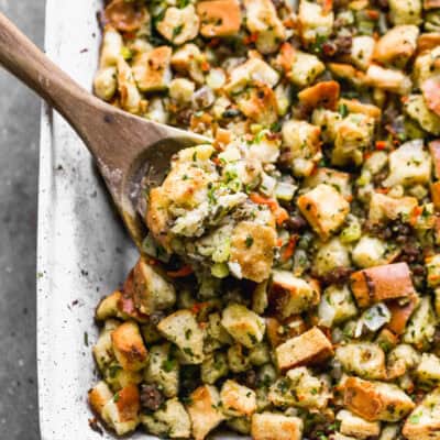 The BEST Thanksgiving Stuffing recipe baked in a white casserole dish with a wooden spoon lifting some up.